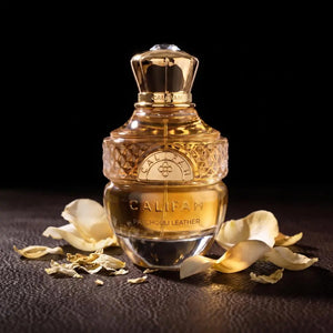 A bottle of Califah Leather Parfum with a textured gold cap and golden liquid inside a clear glass bottle, styled on a dark textured surface with scattered cream-colored flower petals against a black background.