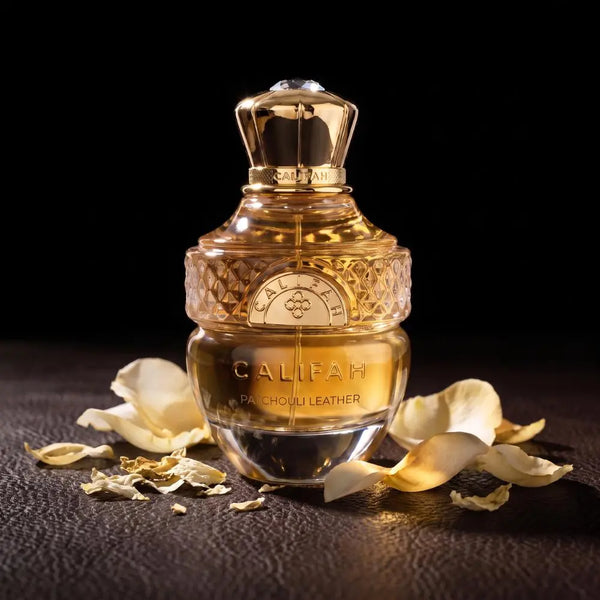 A bottle of Califah Leather Parfum with a textured gold cap and golden liquid inside a clear glass bottle, styled on a dark textured surface with scattered cream-colored flower petals against a black background.