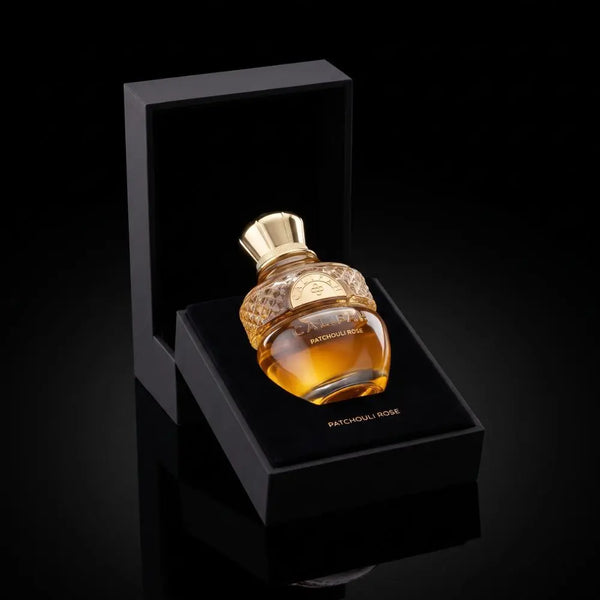 A bottle of Patchouli Rose Parfum with amber liquid and a gold cap, displayed in an open black presentation box on a glossy black background.